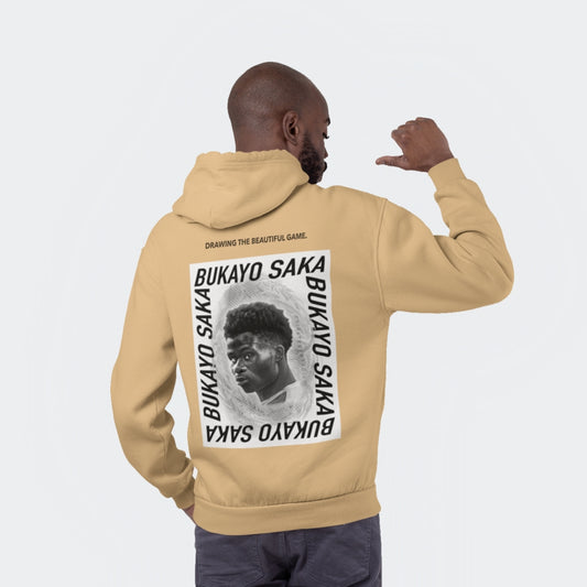 Studio shot of a black male model facing away from the camera wearing a regular fit nude colour hoodie - resembling the shade of sand. On the back of the hoodie there is a large graphic, black and white design featuring a drawing of Arsenal's Bukayo Saka in the centre. Above the large graphic is the lettering 'drawing the beautiful game', which is Goalartzo's slogan.