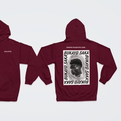 Burgundy Arsenal Bukayo Saka hoodie by Goalartzo laying flat against a pale background. You can see both sides of the hoodie laid flat. There is a logo on the front chest reading 'goalartzo' and a large black and white graphic design featuring Bukayo Saka Art on the back.