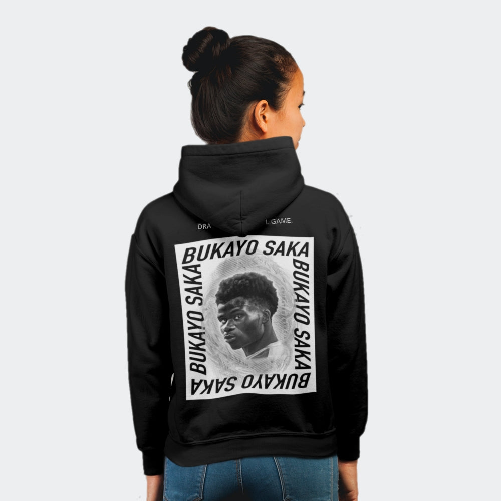 Female model in a photo studio standing facing away from the camera. She wears a black hoodie featuring a large black and white print of Arsenal winger, Bukayo Saka.