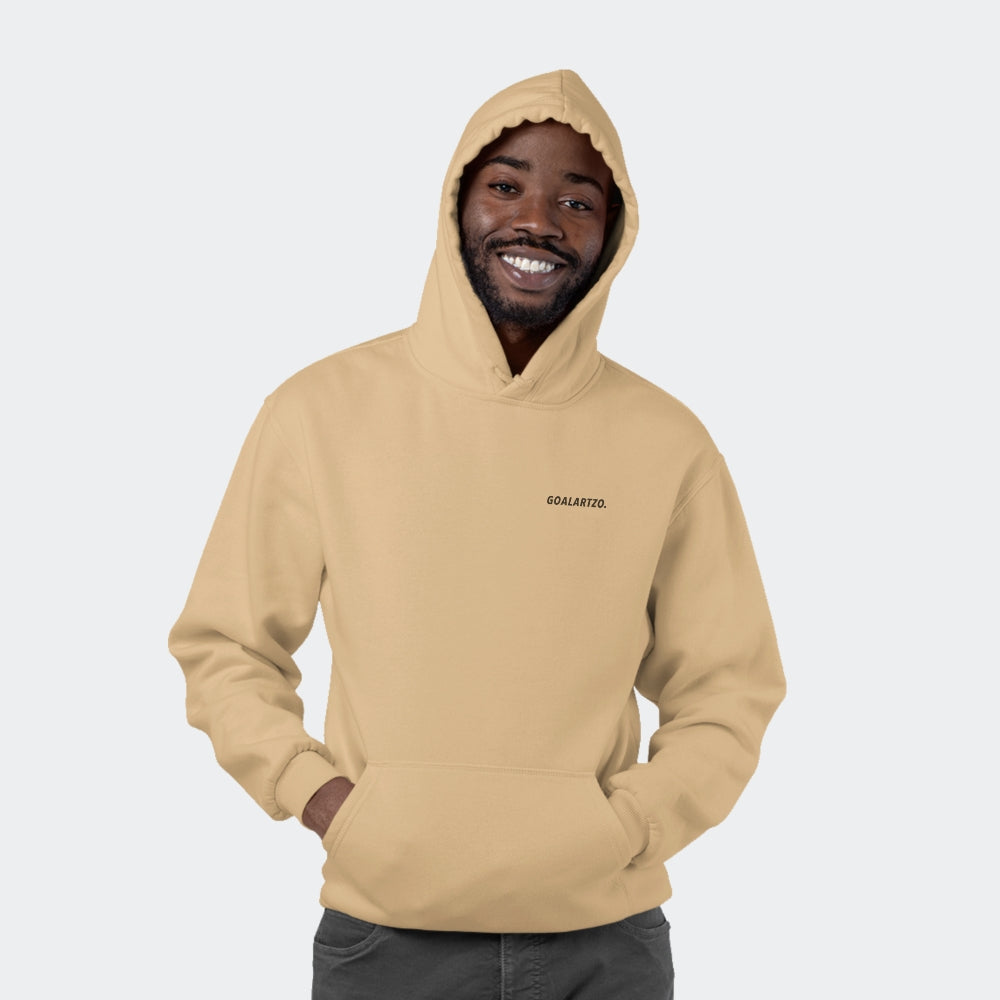 Black male model smiling and facing the camera in a photo studio. He is wearing a sand-coloured nude hoodie with a small logo on his left chest, reading 'Goalartzo.'