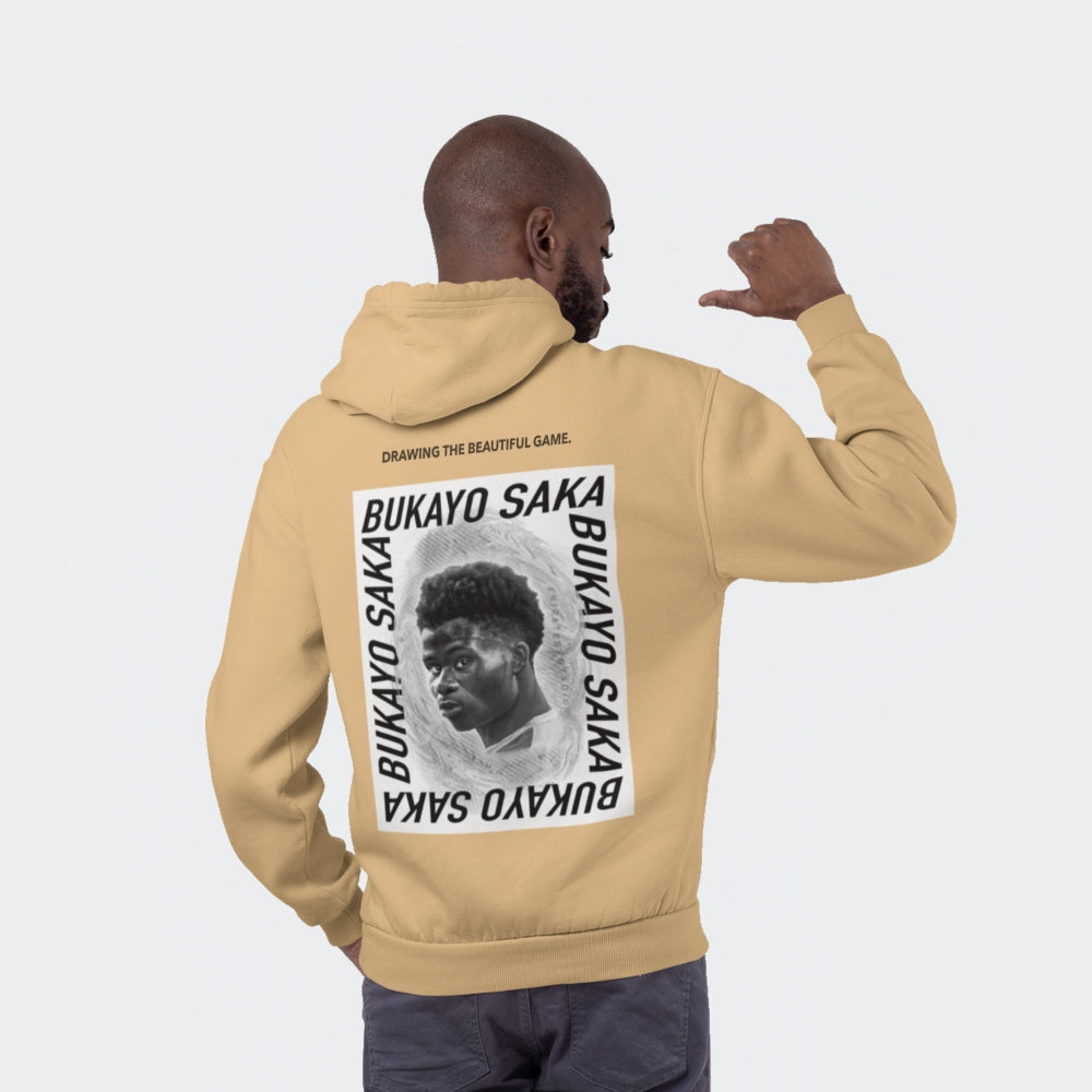 Studio shot of a black male model facing away from the camera wearing a regular fit nude colour hoodie - resembling the shade of sand. On the back of the hoodie there is a large graphic, black and white design featuring a drawing of Arsenal's Bukayo Saka in the centre. Above the large graphic is the lettering 'drawing the beautiful game', which is Goalartzo's slogan.